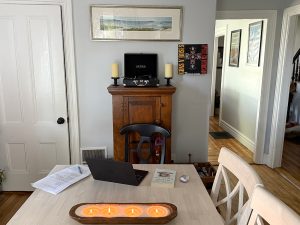A whitewashed table with a candle with four lit wicks, a laptop, a book and a notebook and papers is shown. Behind it is a brown cabinet with a record player. On the wall hangs a beach scene painting and a record - Guns N Roses, Appetite for Destruction.